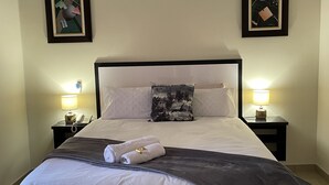 Un lit bien fait, avec du linge de lit blanc et un plaid gris, deux lampes de chevet, un ventilateur de plafond et deux tableaux abstraits accrochés aux murs.