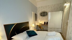 A neatly made bed with white linens, a dark headboard, and a teal pillow. There is a wooden nightstand with a lamp beside the bed. A small desk with a chair and a lamp is visible in the background. - Apartment in Verdun, "La Scène du 66" (Verdun)