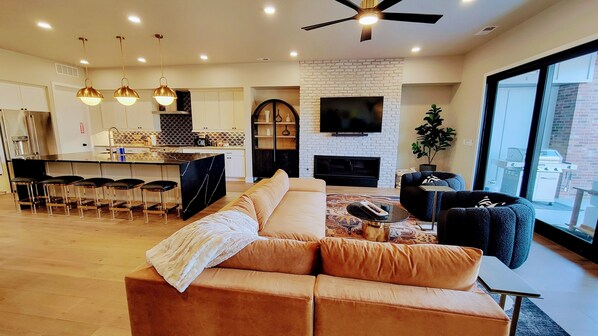 A modern living room with a sectional sofa, a dining area with bar stools, a fireplace, and a large TV mounted on a brick wall.