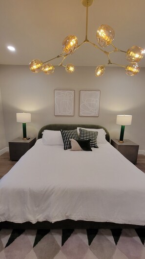 A modern bedroom with a large bed, two bedside tables with lamps, a chandelier, and abstract wall art.