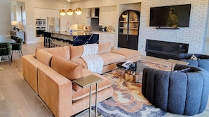 Una sala de estar moderna con un sofá seccional color beige, un sillón azul y una mesa de centro. Se ve una cocina al fondo con gabinetes blancos y electrodomésticos de acero inoxidable.