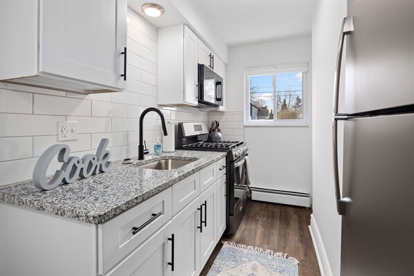 Una cocina con armarios blancos, encimera de granito, refrigerador de acero inoxidable y ventana con vistas al exterior.