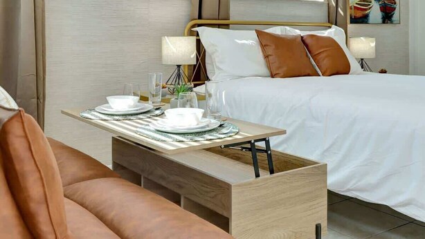Habitación de hotel con cama, mesitas de noche, una pequeña mesa dispuesta con platos y vasos, y una ventana con vistas.