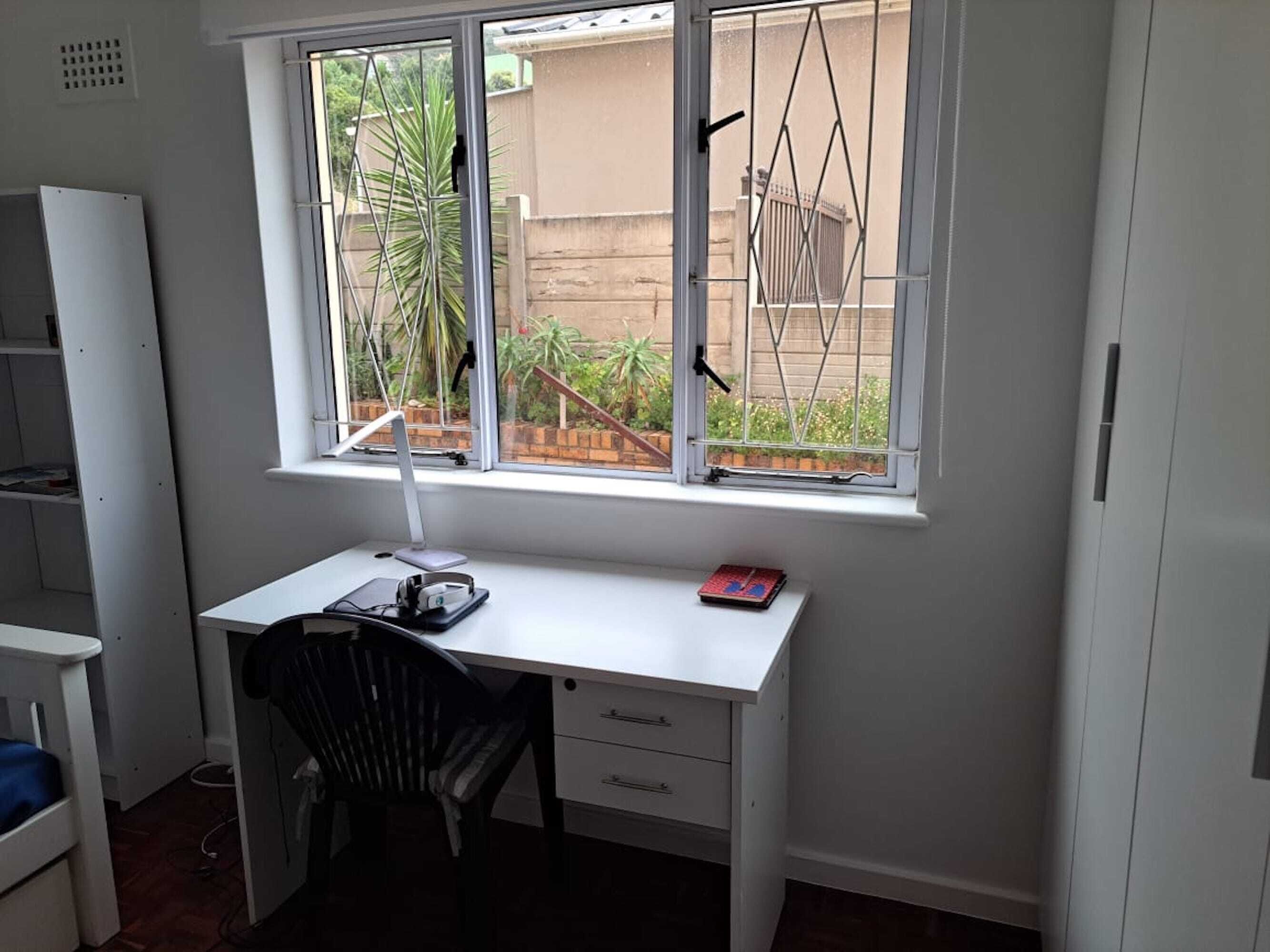 Un pequeño escritorio blanco con una computadora y un libro, ubicado junto a una ventana que da a un jardín y un edificio.