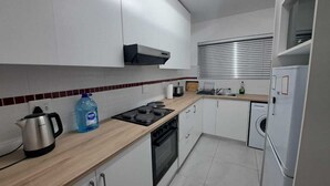 Una cocina compacta con armarios blancos, una tetera de acero inoxidable, un dispensador de agua grande, una estufa, una lavadora y una ventana con persianas.