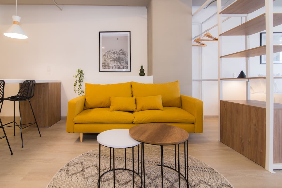 Een moderne woonkamer met een gele bank, een klein wit tafeltje en een rond houten tafeltje. Een zwarte stoel, een plant en een ingelijst foto op de muur.