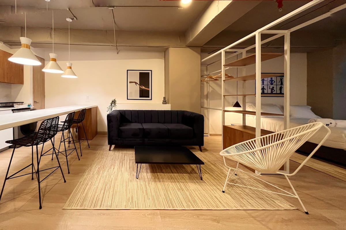 Een moderne woonkamer met een zwarte bank, een witte stoel, een salontafel en een keukenhoek met barkrukken.