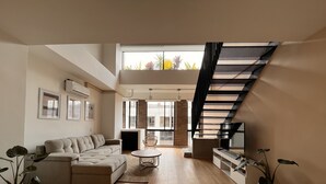 Ein geräumiges Wohnzimmer mit einem beige gefärbten Sofa, einem Couchtisch und Topfpflanzen. Eine Treppe mit schwarzer Geländer führt in einen oberen Bereich.