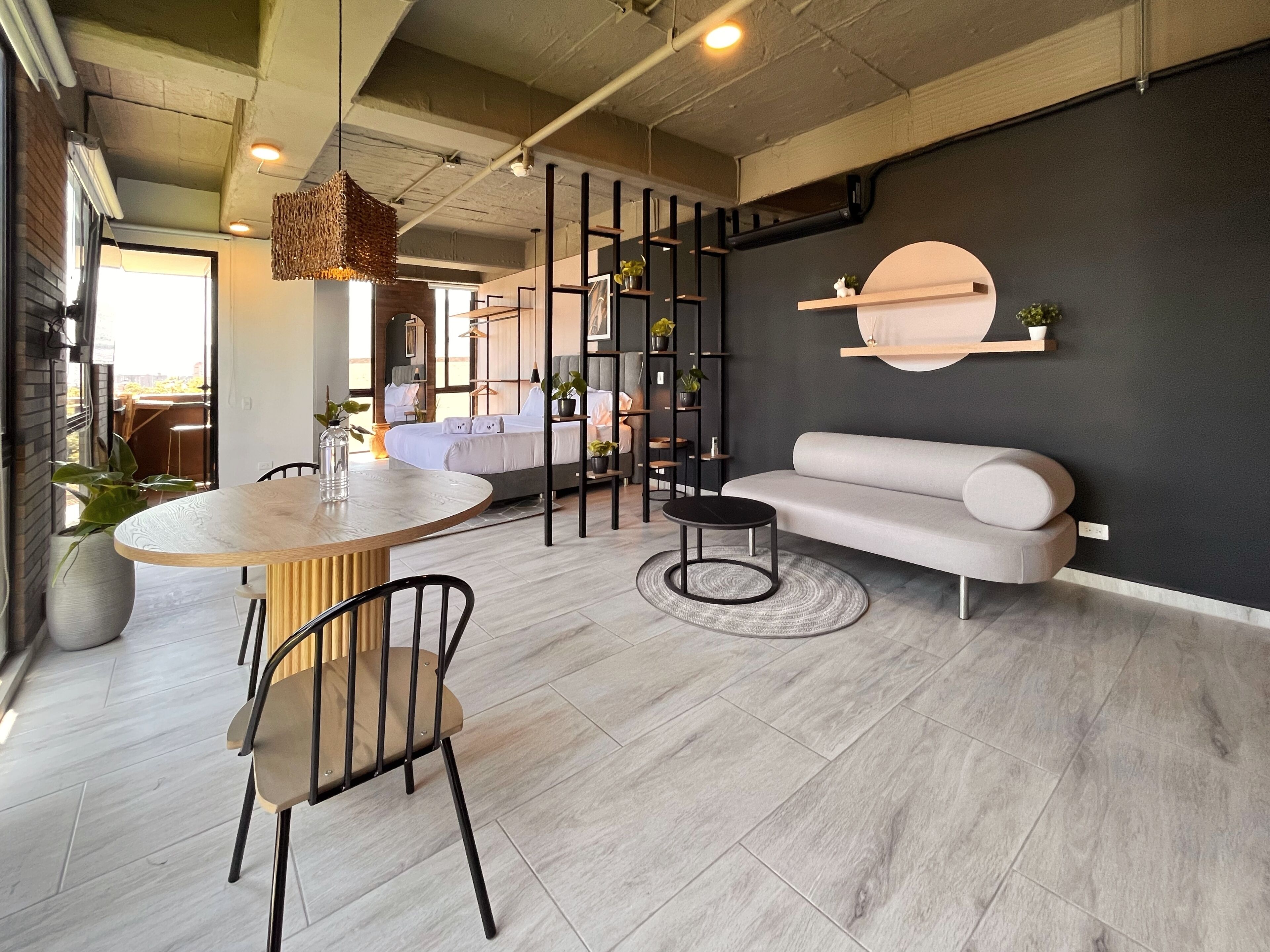 A modern living room with a round dining table, a sofa, and a coffee table. There is a staircase leading to an upper level, and the room has a black accent wall with a round mirror.