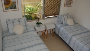 A bedroom with two beds, a small table, and a window with a view of plants and a fence. - Alice Street 1, Unit 1, Currimundi (Currimundi)