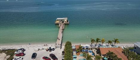 A resort with a pier extending into the ocean, a parking lot, and a pool area with umbrellas.