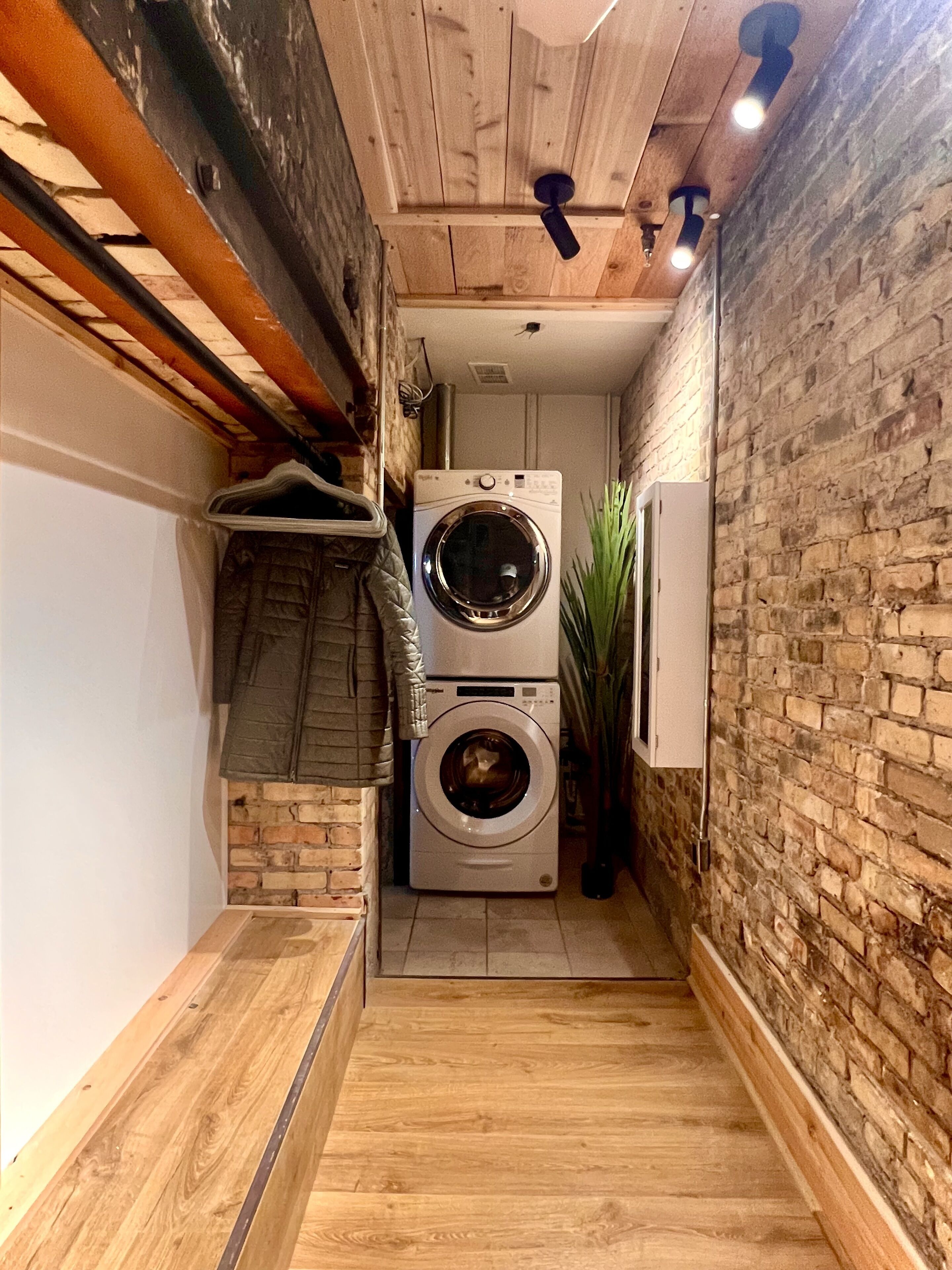 A laundry room with a washing machine and dryer, wooden flooring, a brick wall, and a ceiling with exposed beams and lighting fixtures.