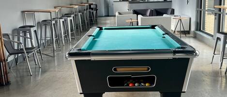 Una mesa de billar en una habitación con techos altos, ventanas amplias y luminarias modernas.