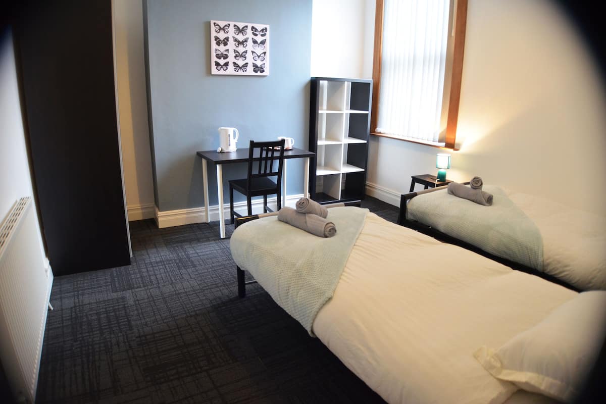 A hotel room with two beds, a desk, a chair, a bookshelf, and a window with blinds.