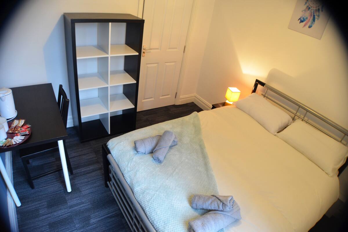 A neatly made bed with pillows and rolled towels, a small table with a cup and snacks, and a wardrobe with shelves.