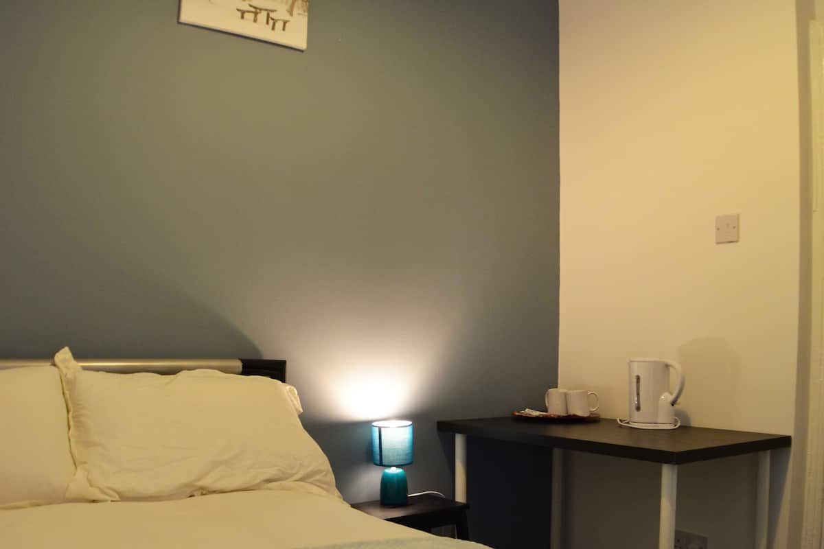 A neatly made bed with a rolled towel, a bedside table with a lamp, and a small table with a cup and a kettle.