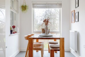 A bright room with a wooden table, chairs, a vase with dried flowers, and a window with blinds. - Central & Modern 1BD Flat with Balcony, Hackney! (London)