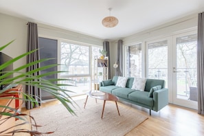 A spacious living room with a green sofa, a coffee table, and a large window offering a view of the outdoors. - Central & Modern 1BD Flat with Balcony, Hackney! (London)