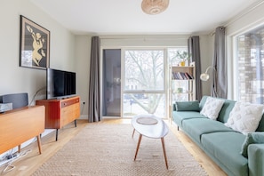 A living room with a green sofa, a small white table, a TV on a wooden cabinet, and a bookshelf. - Central & Modern 1BD Flat with Balcony, Hackney! (London)