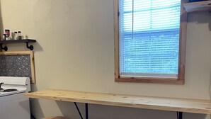 Eine Küche mit einer Holzarbeitsplatte, drei hölzernen Barhocker, einem Fenster mit Jalousien und einer weißen Mülltonne.