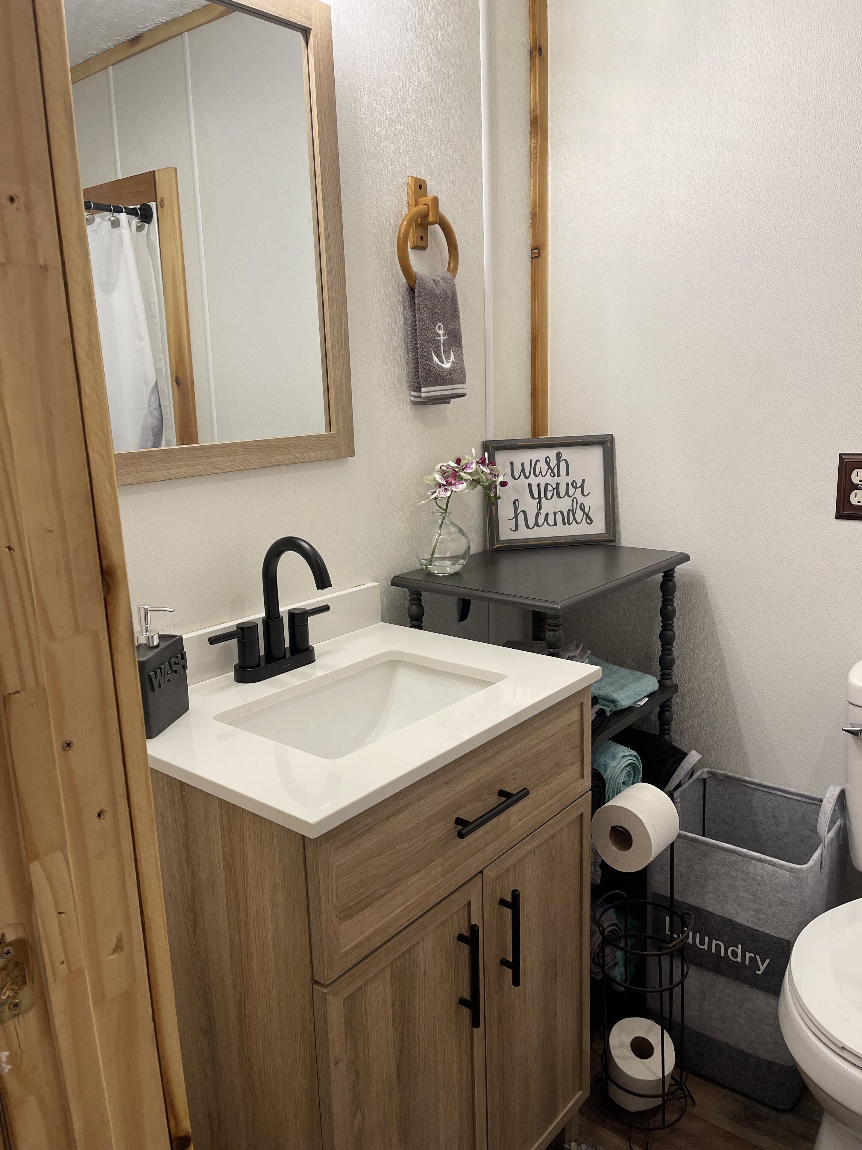A bathroom with a wooden vanity, a sink, a mirror, a towel rack, a framed sign, and a laundry basket.