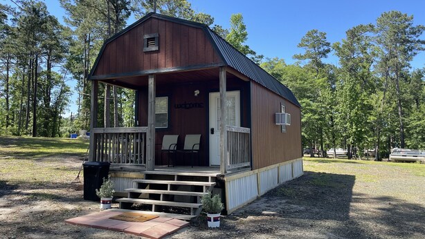 Eine kleine Holzhütte mit einer Veranda, einem Willkommensschild und ein paar Stühlen davor.