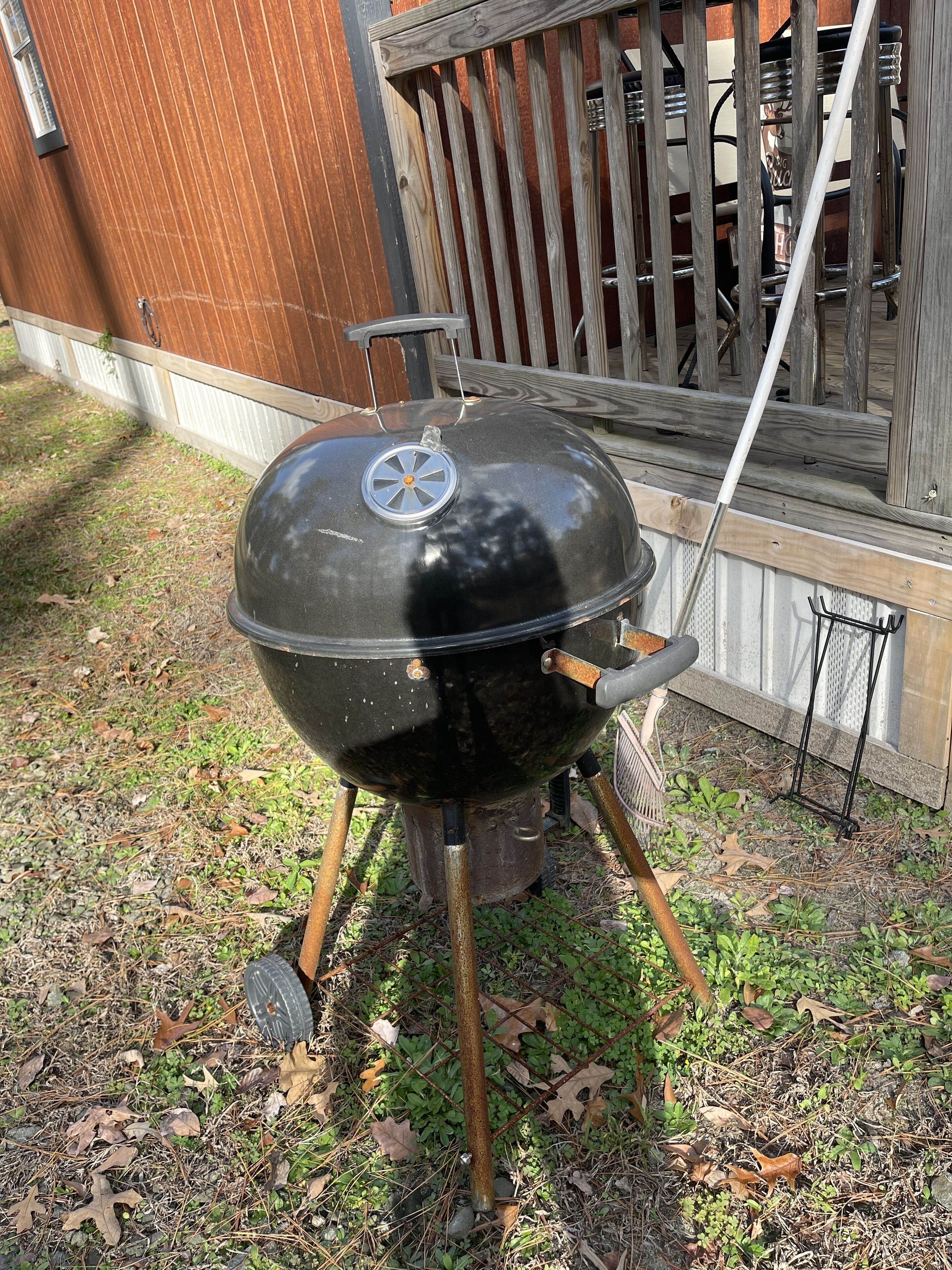 A black barbecue grill with a lid, placed outdoors on a grassy area with a wooden fence and a window in the background.