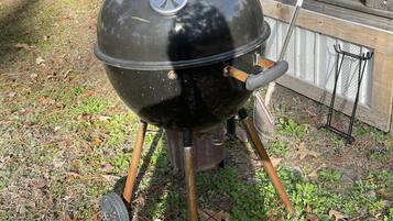 A black barbecue grill with a lid, placed outdoors on a grassy area with a wooden fence and a window in the background.