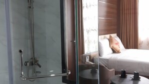 A modern bathroom with a glass shower enclosure, a bed with white linens and a brown pillow, and a small round table with two cups. - palermohotel  (Hanoi)