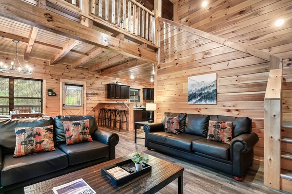 Ein gemütliches Blockhauss-Interieur mit Holzwänden, zwei schwarzen Ledersofas mit gemusterten Kissen, einem Holztisch und einem Gemälde an der Wand.