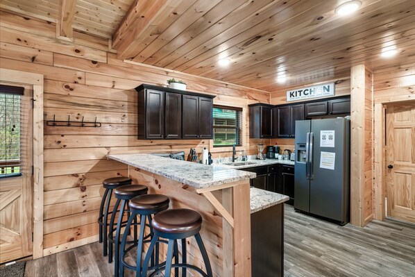 A wooden cabin interior with a kitchen area featuring dark cabinets, a granite countertop, and a refrigerator. - 1mi to PF Prkwy! Hot Tub + Pool Table + Arcade (Pigeon Forge)