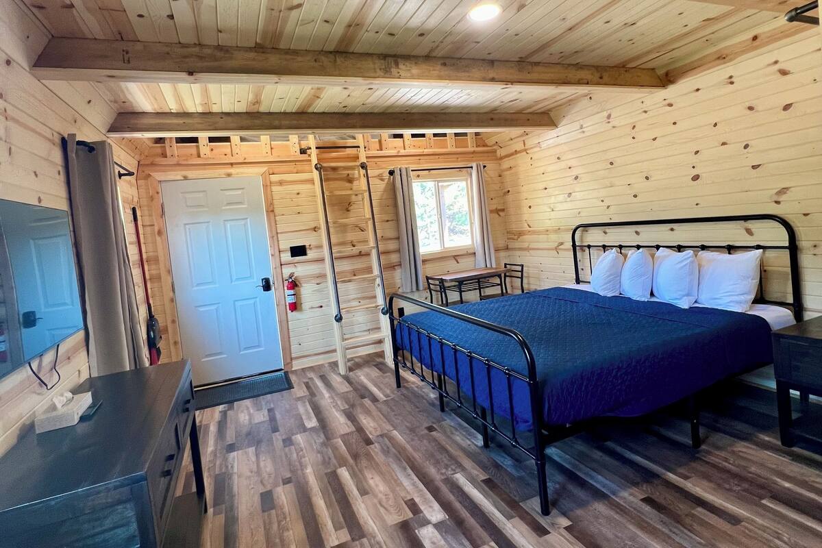 A wooden cabin interior with a bed, desk, and a door leading to another room.