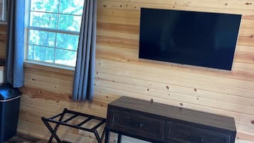 A cozy cabin interior with a wooden ceiling, a large window with curtains, a flat-screen TV mounted on the wall, and a dark wooden bench with drawers.