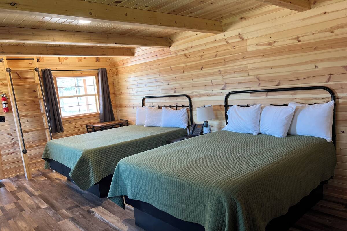 A cabin-style room with two beds, wooden walls, a window with curtains, a wooden ladder, and a small table.