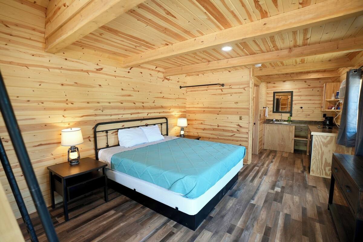A wooden cabin bedroom with a bed, bedside tables, a small kitchenette, and a wooden ceiling.