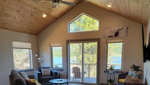 A spacious living area with a high ceiling, wooden beams, a ceiling fan, and large windows offering views of a lake and trees. - Lakeshore Cabins (Homer)