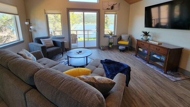 A spacious living room with a large sofa, a coffee table, and a TV mounted on the wall. There is a balcony with a view of a lake. - Lakeshore Cabins (Homer)