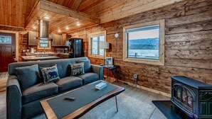 A cozy cabin interior with a wooden interior, a sofa, a coffee table, a fireplace, and a kitchen area. - Lakeshore Cabins (Homer)