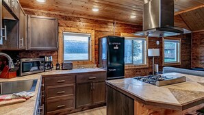 A modern kitchen with wooden walls, a large island, stainless steel appliances, and a view of the outdoors. - Lakeshore Cabins (Homer)