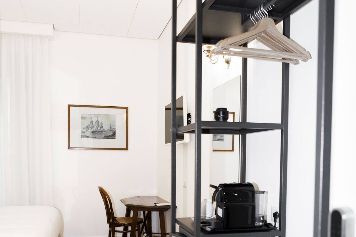 A hotel room with a bed, a dining table with chairs, a coffee machine, and a black shelving unit with a hanging light.