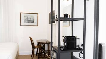 A hotel room with a bed, a dining table with chairs, a coffee machine, and a black shelving unit with a hanging light.