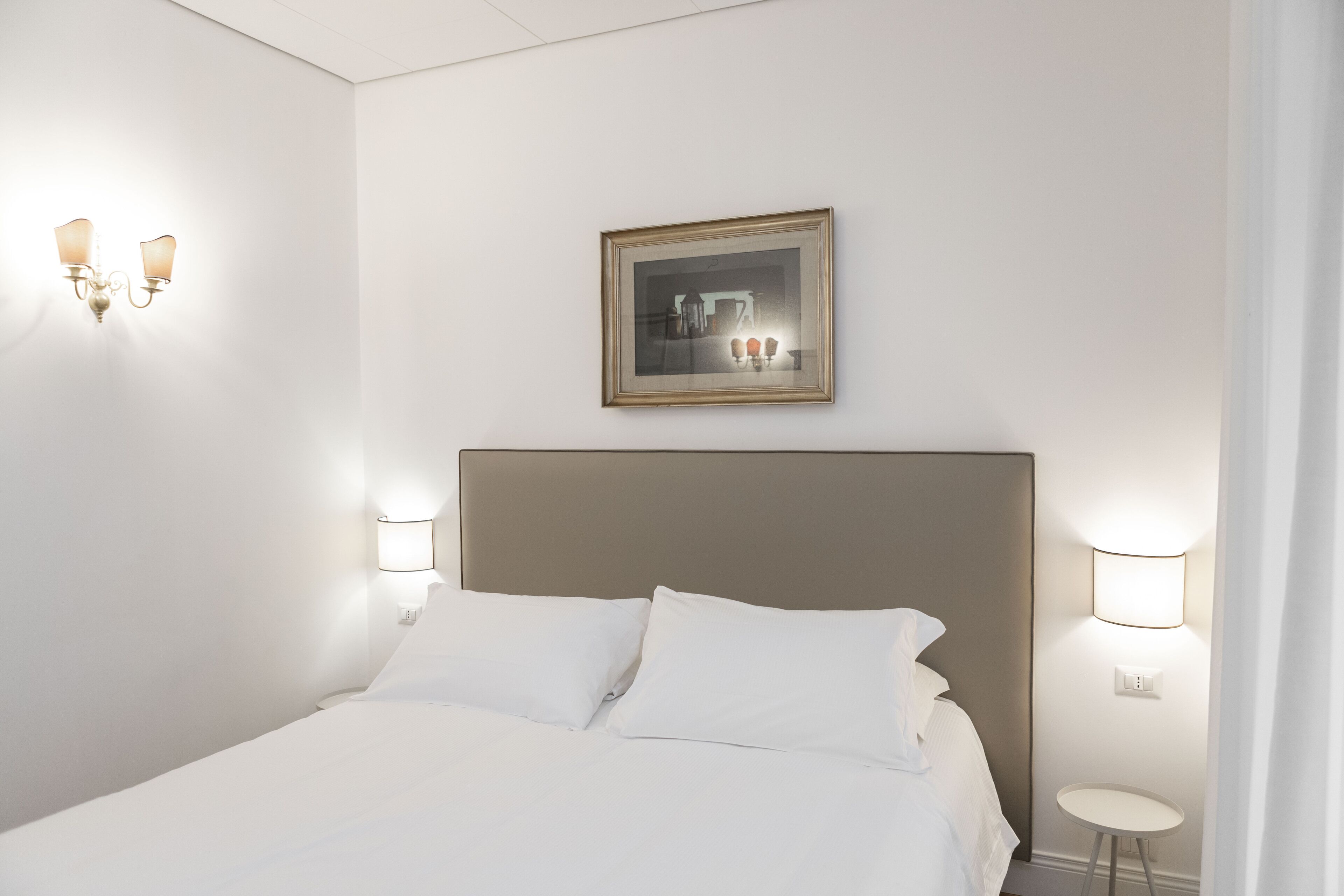 Una cama bien tendida con sábanas blancas, un cabecero y dos lámparas fijadas a la pared.