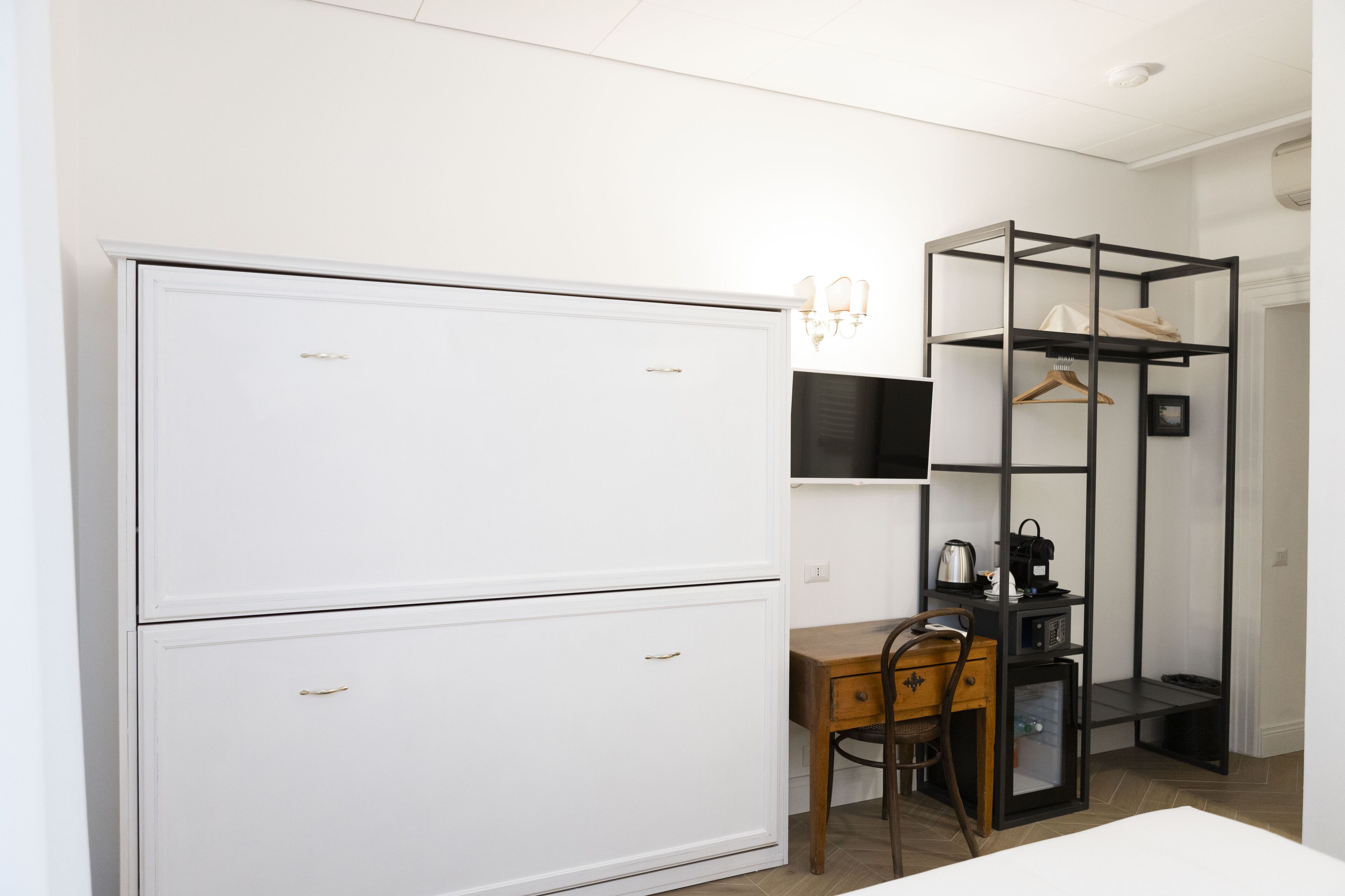 Quarto compacto com guarda-roupa branco, escrivaninha com cadeira, prateleira suspensa e um pequeno armário.