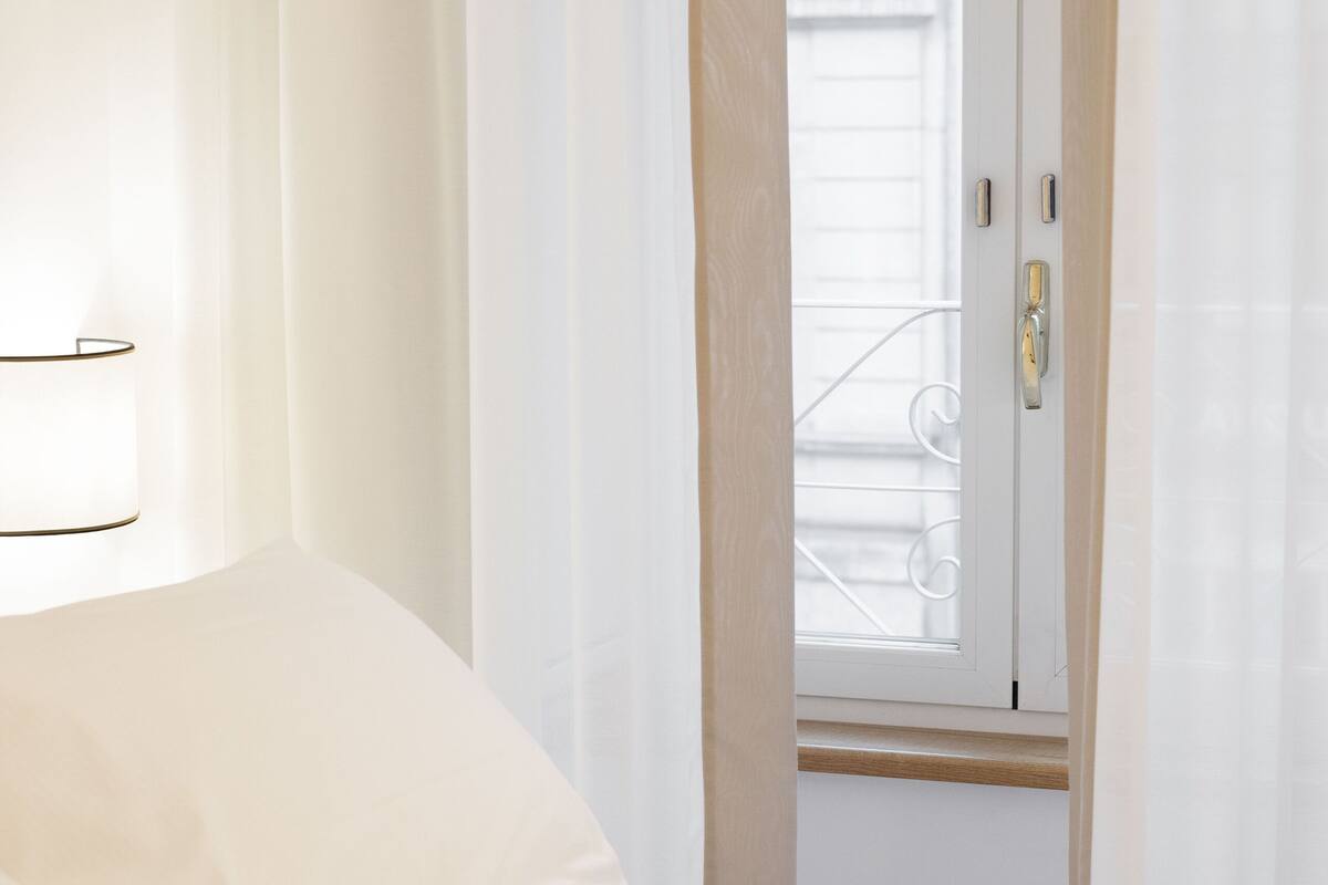 A hotel room with a bed, white curtains, and a window with a metal railing.