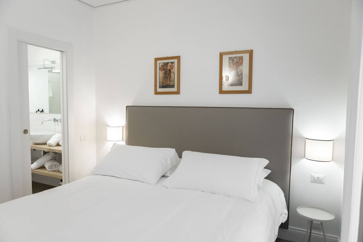A hotel room with a bed, a small bedside table, a wall-mounted light, and a framed picture above the bed.
