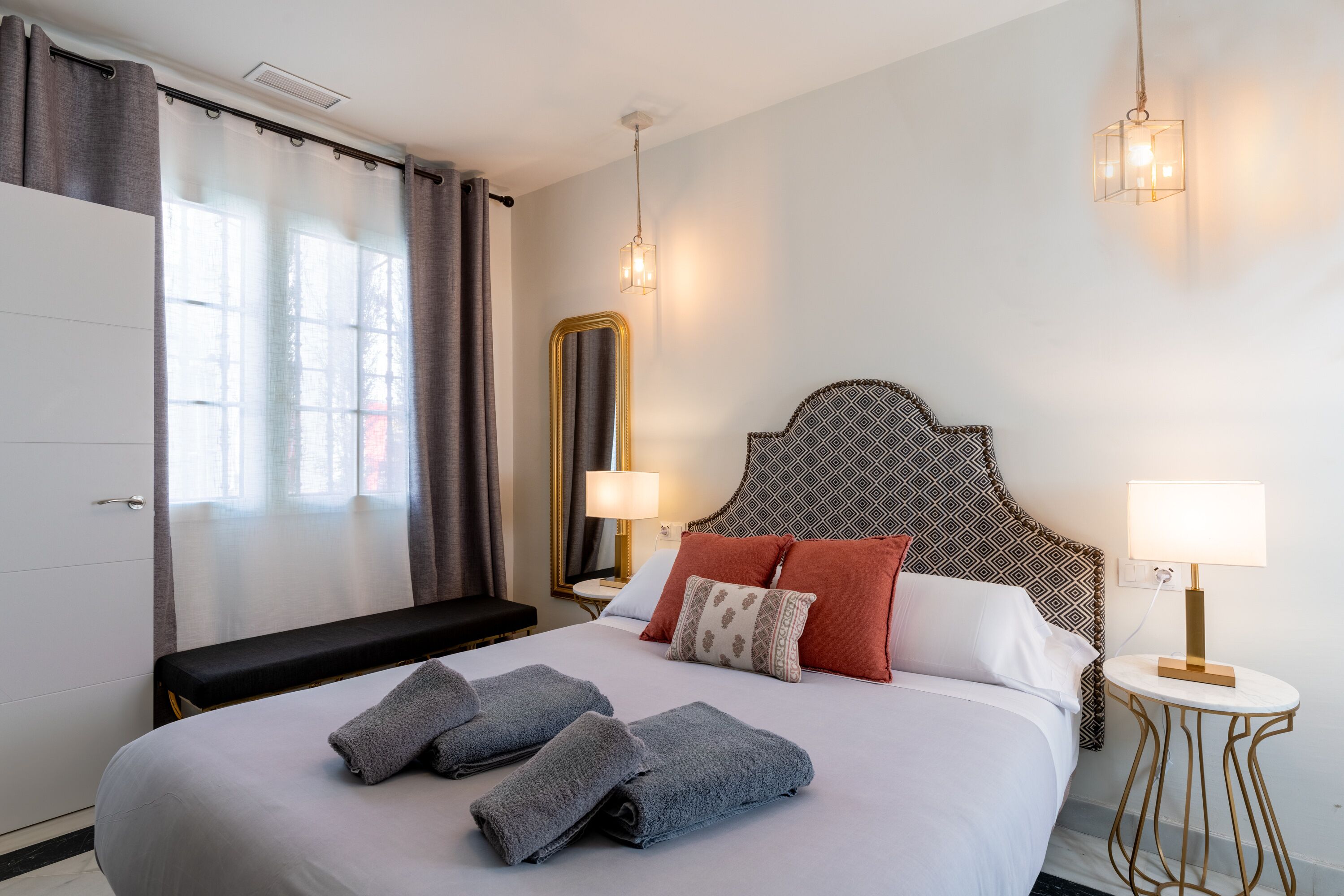 A hotel room with a large bed, two bedside lamps, a bench, and a view of the outdoors through the window.