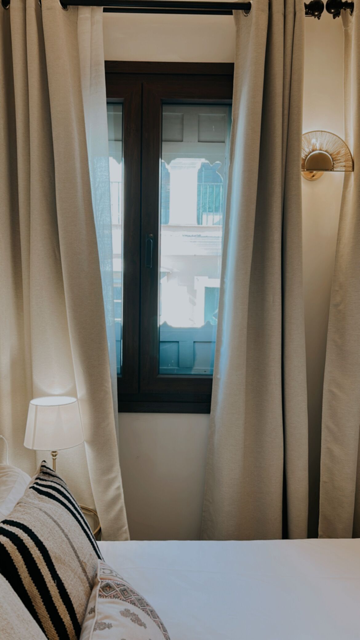 Een slaapkamer met raam, gordijnen, een lamp en een bed met gestreepte kussens.