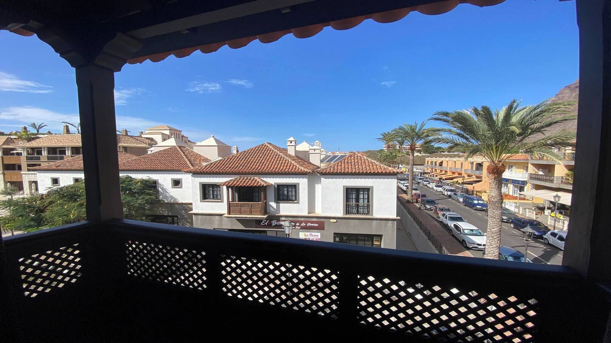 Blick von einem Balkon mit Gittergeländer auf eine Straße mit Gebäuden, Palmen und parkenden Autos.