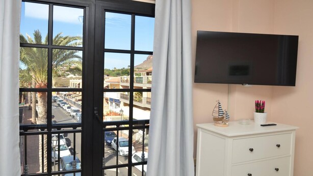 A hotel room with a balcony, a TV mounted on the wall, a small table with decorative items, and a view of a street with palm trees and parked cars. - Apt. Brisa Marina  (Valle Gran Rey)
