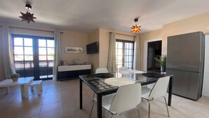 A modern kitchen with a glass dining table, white chairs, a refrigerator, and a TV mounted on the wall. - Apt. Brisa Marina  (Valle Gran Rey)